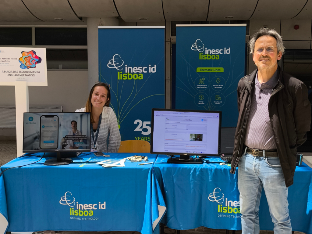 Two researchers standing at the INESC-ID stand at Técnico Oeiras Open Day, with branded banners and demonstration screens visible in the background.