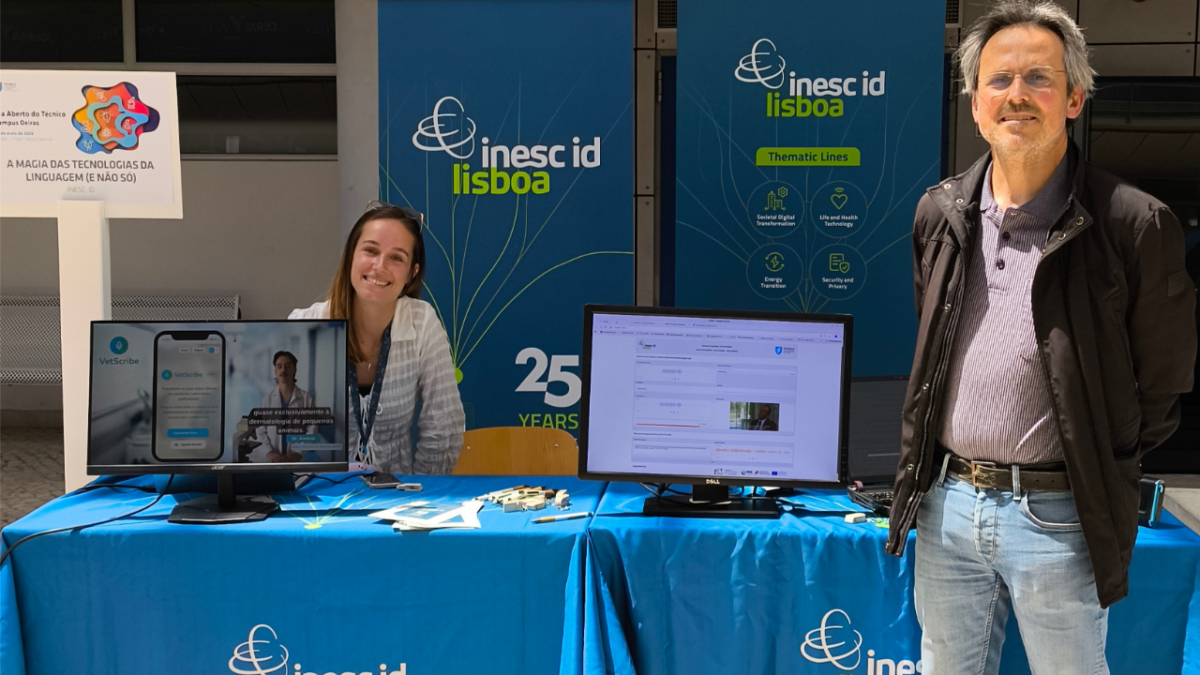 Two researchers standing at the INESC-ID stand at Técnico Oeiras Open Day, with branded banners and demonstration screens visible in the background.